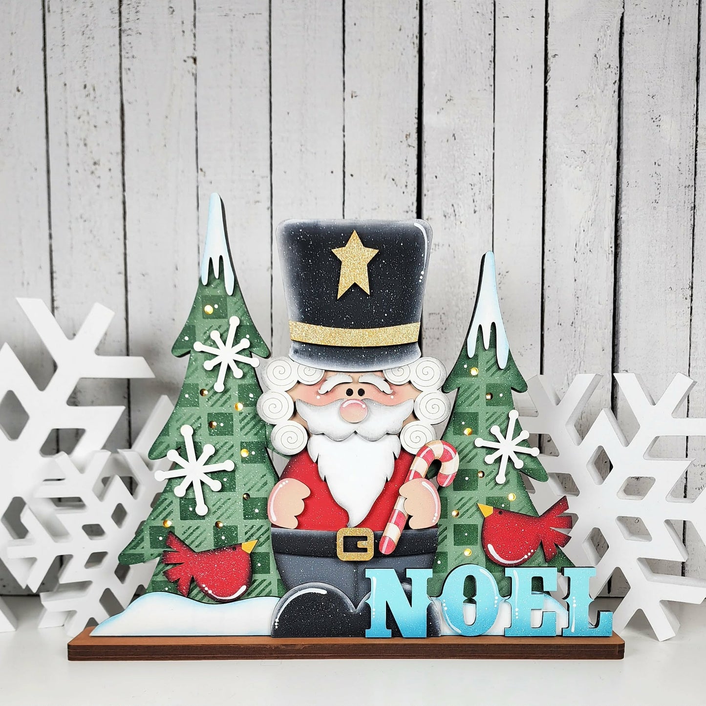 Decorative Christmas scene with a Santa Claus figure, trees, and 'NOEL' text on a wooden background.