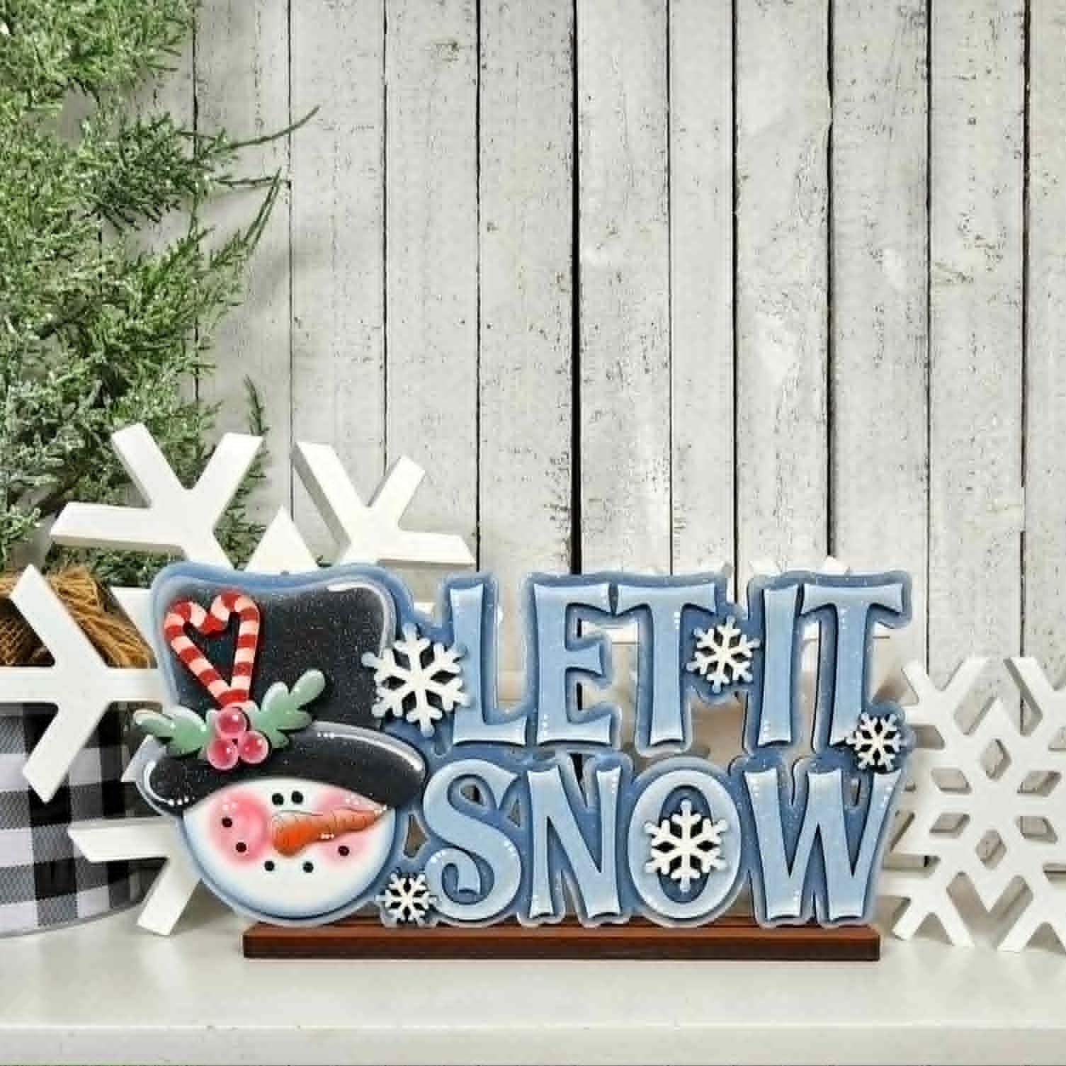 Decorative sign with 'Let It Snow' text and snowman design on a wooden background