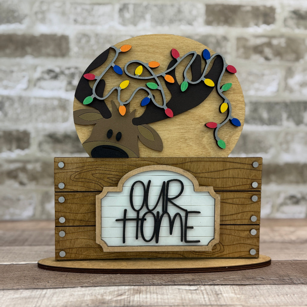 Wooden decorative sign with reindeer, lights, and 'Our Home' text against a brick wall.