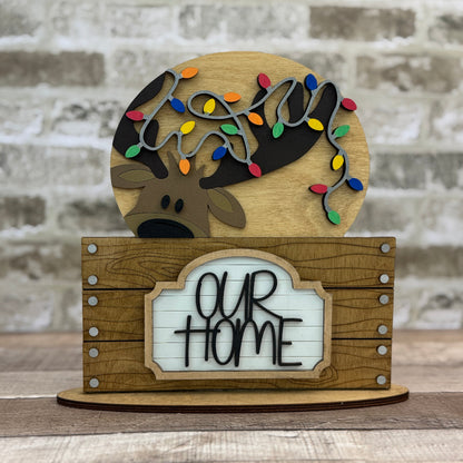 Wooden decorative sign with reindeer, lights, and 'Our Home' text against a brick wall.