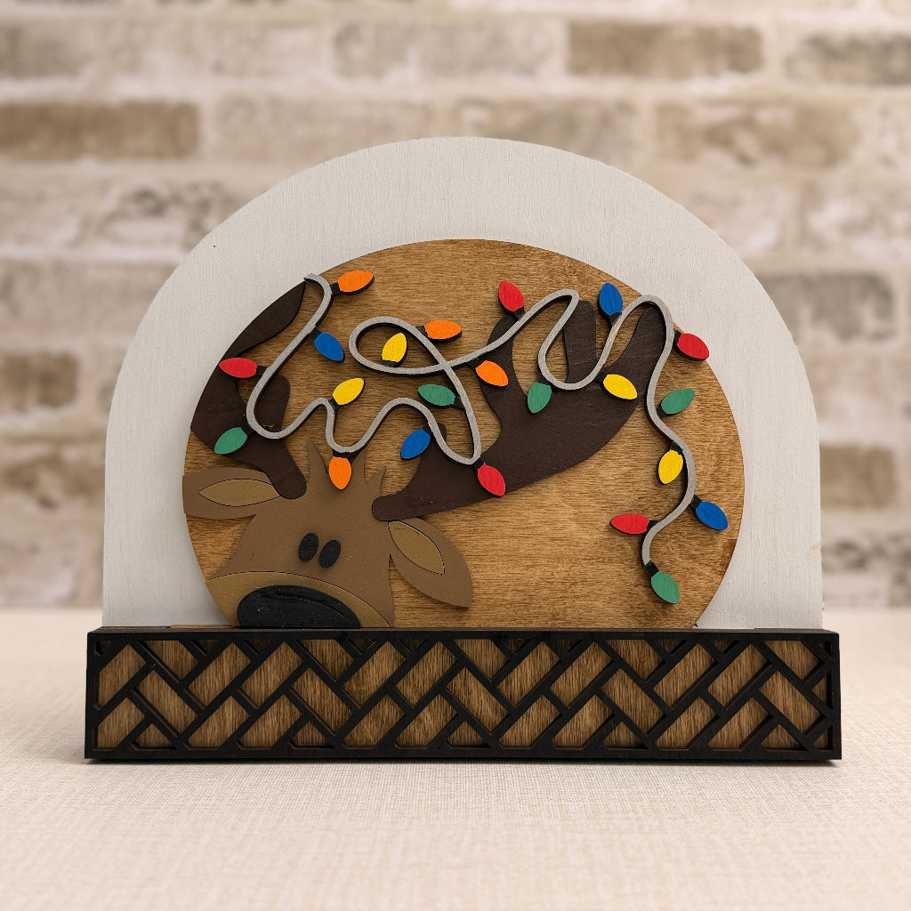 Decorative sign with a reindeer and Christmas lights on a white arch, against a brick wall.