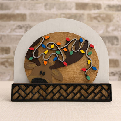 Decorative sign with a reindeer and Christmas lights on a white arch, against a brick wall.