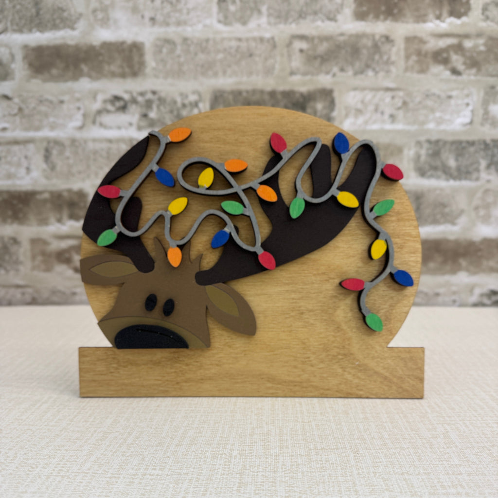 Decorative wooden sign with reindeer and lights on a white surface with brick wall background
