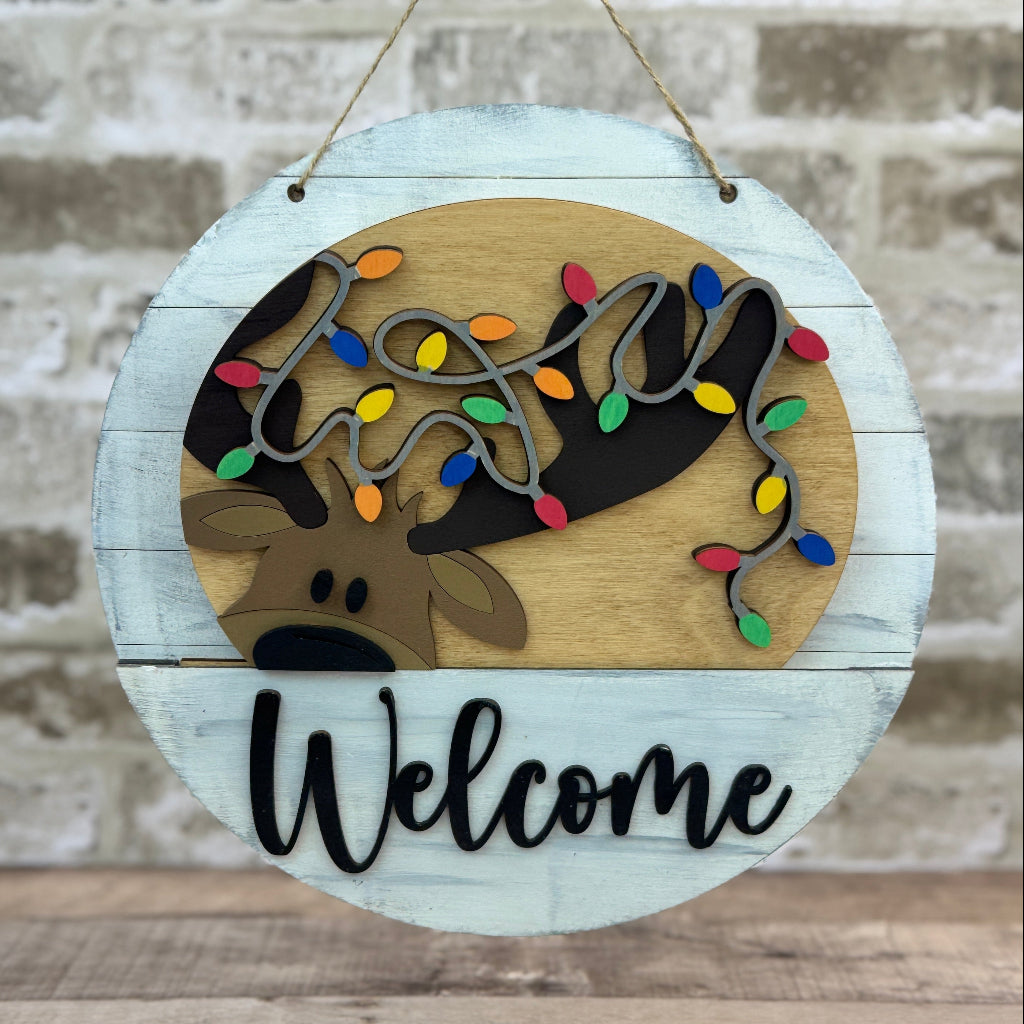 Round wooden sign with reindeer design and 'Welcome' text, hanging against a textured wall.