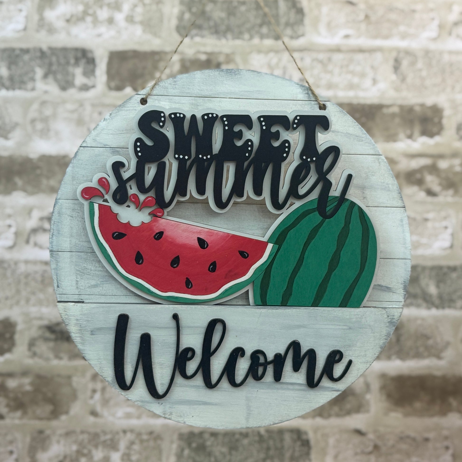 Round wooden sign with 'Sweet Summer Welcome' text and watermelon graphics on a stone wall background.