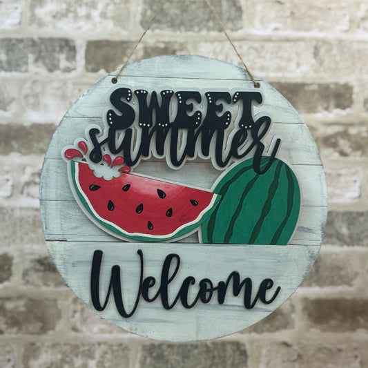 Round wooden sign with 'Sweet Summer Welcome' text and watermelon graphics on a stone wall background.